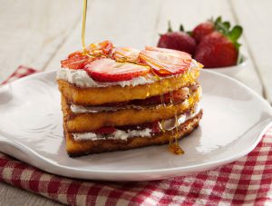 Three heart-shaped French toasts with strawberries on top, and cream on top and in between, on a plate. A bowl of strawberries next to it.