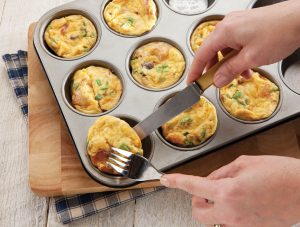Someone using a fork and knife to remove frittata muffins from the mold of the baking tray. Some molds still having muffins in them.